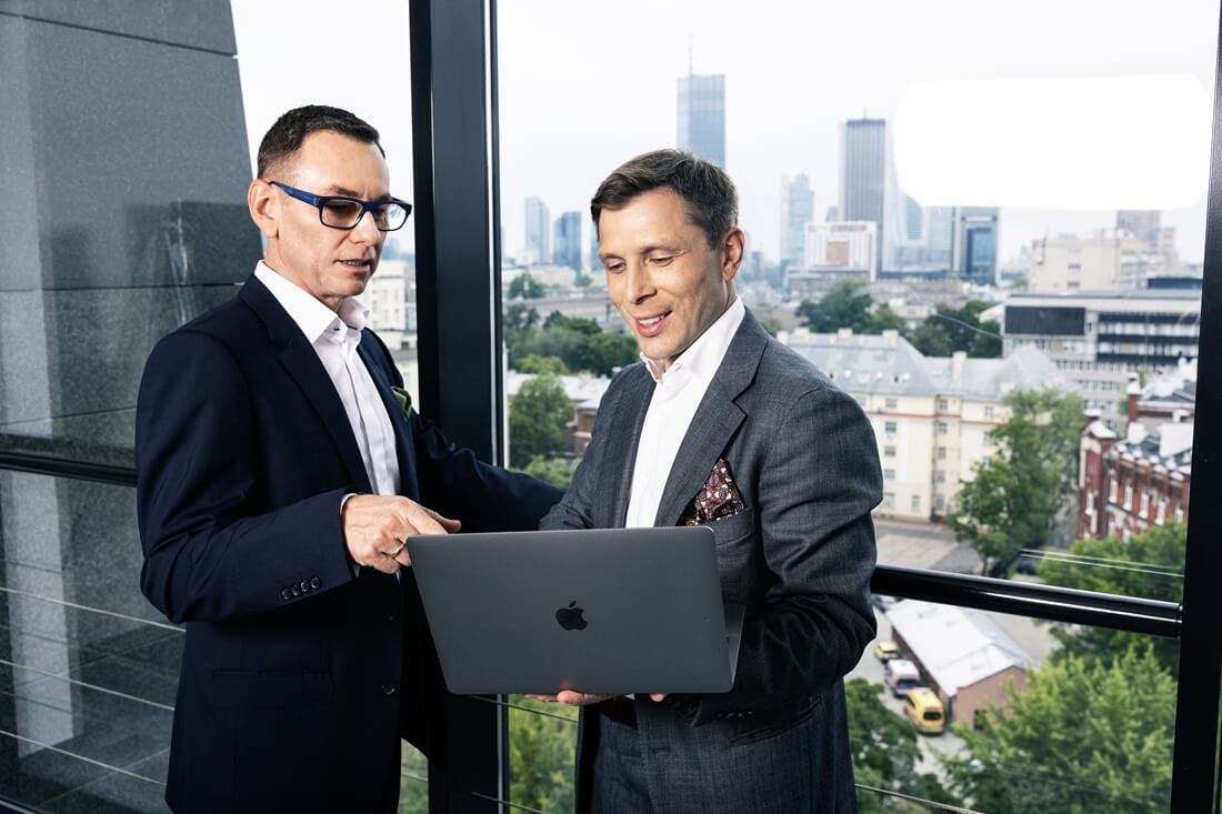 Na zdjęciu widać dwóch elegancko ubranych mężczyzn stojących przy dużym oknie w nowoczesnym biurze. Jeden z nich trzyma laptopa marki Apple, a drugi wskazuje coś na ekranie. Obaj wyglądają na skupionych i prowadzą rozmowę dotyczącą pracy. W tle znajduje się panorama miasta z wysokimi budynkami oraz zielenią.