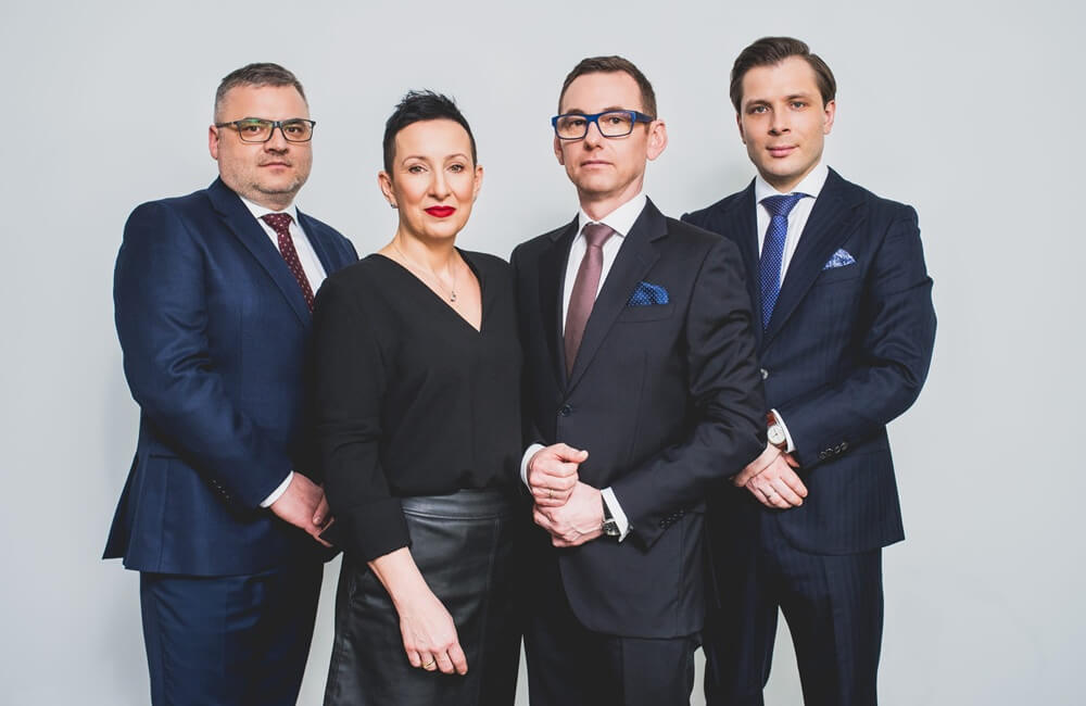 Zdjęcie to formalny portret grupowy, na którym widać cztery osoby — trzech mężczyzn i jedną kobietę — stojących razem na jasnym, jednolitym tle. Wszyscy są ubrani w stroje biznesowe.