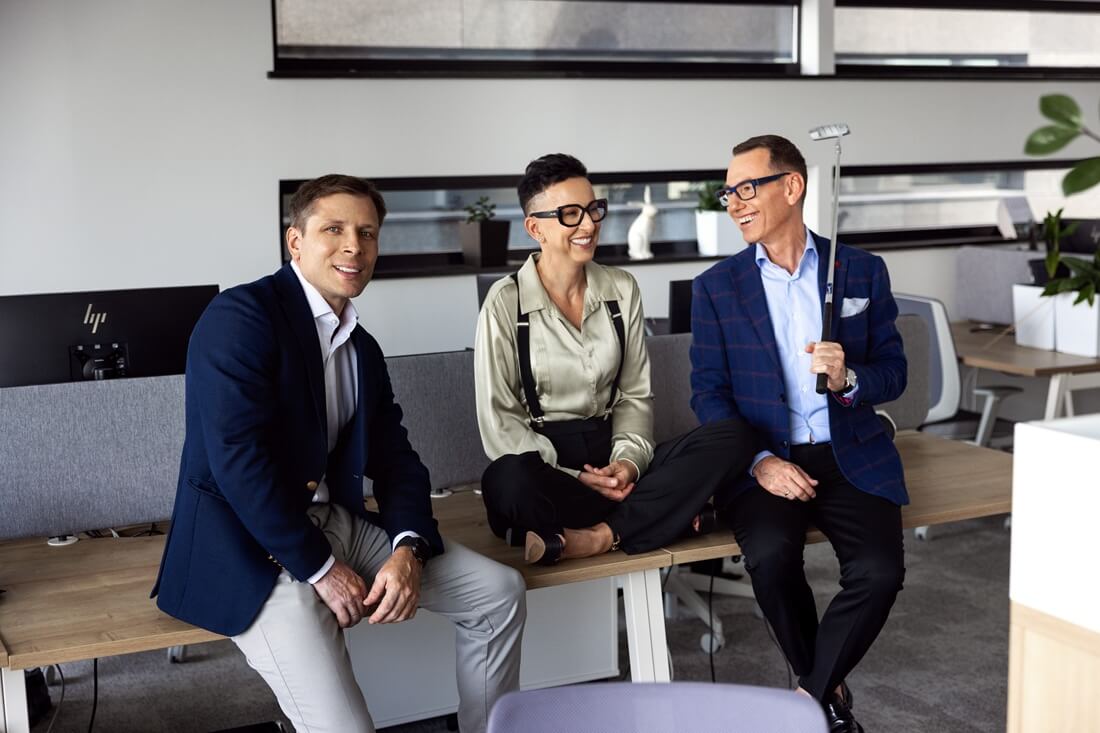 Trzy osoby siedzą na długim, drewnianym blacie w nowoczesnym biurze. Ubrane są w eleganckie stroje - od granatowych marynarek po stylowe dodatki, takie jak okulary czy zegarek, co podkreśla ich biznesowy charakter. Mężczyzna po prawej stronie trzyma kij golfowy. Kobieta pośrodku siedzi po turecku. W tle widać biurowe krzesła, monitory i rośliny, które nadają przestrzeni przyjazny, funkcjonalny klimat. 