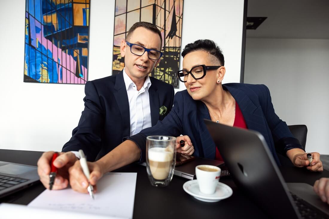 Na zdjęciu widać dwie osoby w eleganckich, biznesowych strojach siedzące przy biurku w nowoczesnym biurze. Oboje mają na sobie granatowe marynarki i okulary, a przed nimi stoją laptopy, filiżanki kawy oraz dokumenty. Kobieta i mężczyzna wspólnie analizują notatki — on trzyma długopis i pokazuje coś na kartce, a ona z zainteresowaniem się temu przygląda. W tle widoczne są kolorowe obrazy o geometrycznych wzorach, które nadają wnętrzu nowoczesny charakter.
