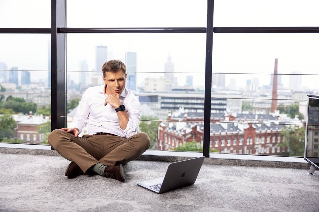 Na zdjęciu widać mężczyznę siedzącego na podłodze w biurze z panoramicznym widokiem na miasto. Przed nim stoi otwarty laptop, w który uważnie spogląda, wspierając brodę na dłoni w zamyśleniu. Ma na sobie białą koszulę i brązowe spodnie, a za jego plecami widać duże okna z widokiem na warszawskie wieżowce i zabudowę miejską. Scena oddaje atmosferę skupienia i pracy w nowoczesnym, miejskim otoczeniu — symbolizuje elastyczność, nowoczesne podejście do pracy i niezależność zawodową.