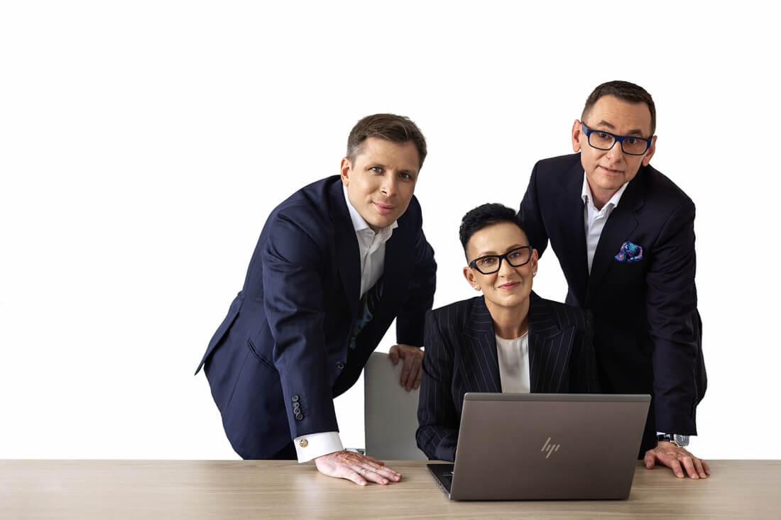 Na zdjęciu widzimy trzy osoby w eleganckich strojach biznesowych zgromadzone wokół laptopa ustawionego na drewnianym stole. Jedna z nich siedzi bezpośrednio przed komputerem, a dwie pozostałe stoją po bokach — wszyscy patrzą w stronę aparatu. Tło jest całkowicie białe, co nadaje scenie profesjonalny, minimalistyczny charakter.