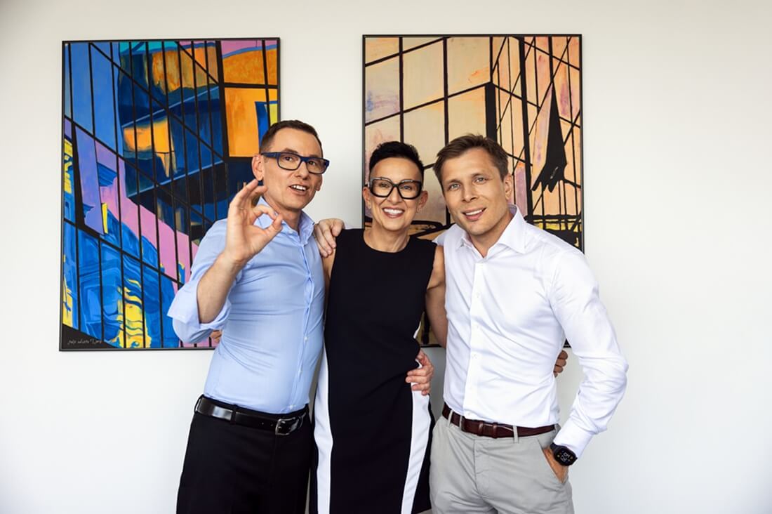 Na zdjęciu widzimy trzy osoby stojące razem na tle białej ściany, ozdobionej dwoma kolorowymi obrazami o abstrakcyjnych, geometrycznych wzorach. Mężczyzna po lewej, w jasnoniebieskiej koszuli i okularach, pokazuje gest „OK” dłonią. W środku stoi uśmiechnięta kobieta w czarno-białej sukience bez rękawów, obejmująca pozostałe osoby. Po prawej znajduje się mężczyzna w białej koszuli, z zegarkiem na lewym nadgarstku.