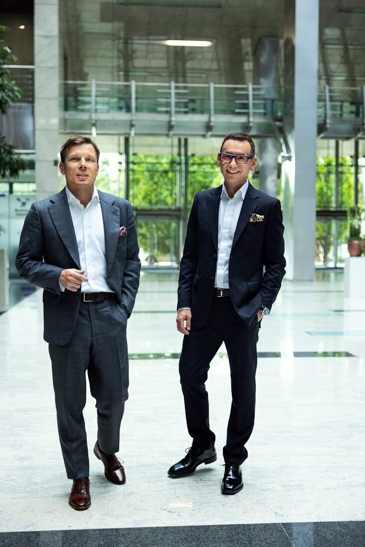 Na zdjęciu widzimy dwóch uśmiechniętych mężczyzn w eleganckich garniturach stojących w przestronnym, nowoczesnym wnętrzu. Jeden ubrany jest w jasnoszary garnitur z białą koszulą i brązowymi butami, drugi w ciemnogranatowy lub czarny garnitur z czarnymi butami. Tło tworzy jasna podłoga, duże przeszklenia wpuszczające naturalne światło oraz elementy architektoniczne — metalowe balustrady i rośliny doniczkowe.